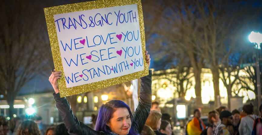 Trans Rights Are Human Rights - A demonstrator with purple hair holds a protest sign above their head that reads "Trans & GNC Youth - We We Love You - We See You - We Stand With You"